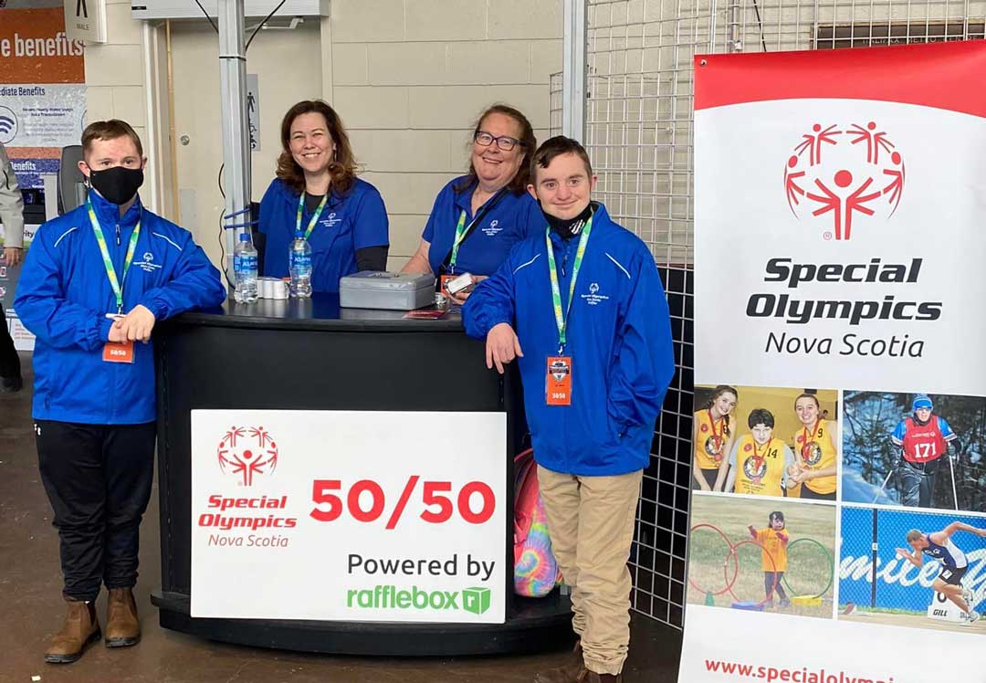 Four volunteers in blue Special Olympics Nova Scotia jackets standing behind a booth with a 50/50 raffle sign powered by Rafflebox. A Special Olympics Nova Scotia banner is visible to the right displaying athletes participating in various sports.