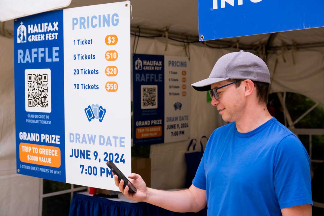 Man in blue t-shirt and gray cap scanning a QR code on a Halifax Greek Fest raffle sign. The sign shows pricing options ($3 for 1 ticket up to $50 for 70 tickets) and advertises a grand prize trip to Greece valued at $3000, with draw date June 9, 2024 at 7:00 PM.
