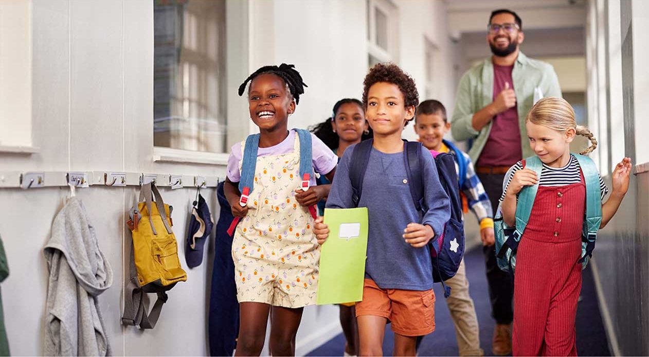Group of diverse children walking in a school hallway with a teacher. Students wearing colorful clothing and backpacks, with the front child holding a green folder. Coat hooks with jackets visible on the wall.